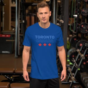 Op de afbeelding: Een koningsblauw t-shirt met korte mouwen voorzien van de tekst TORONTO CANADA in een lichtblauw lettertype, geplaatst boven drie kleine, effen rode esdoornbladpictogrammen in het midden op de borst.