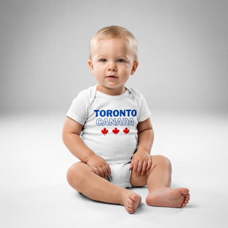 以下が含まれることがあります： 白い半袖のベビー用ボディスーツ。胸元中央に青い文字で「TORONTO」、白い縁取りの文字で「CANADA」とプリントされ、その下に3つの小さな赤いカエデの葉のアイコンが並んでいます。クラシックなクルーネックのデザインです。