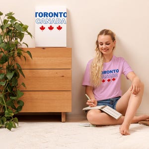 Op de afbeelding: Een lichtpaars t-shirt met de tekst TORONTO CANADA in blauwe en witte letters, vergezeld van drie rode esdoornblad-iconen. Het ontwerp is op de borst van het shirt gedrukt en komt overeen met de afbeelding op de witte wanddecoratie.