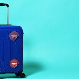 12 Canada cities as glitter pin badges - transparent PNG + printable A4 sheet. Print at home on sticker paper, cut, and use for journals, laptops, water bottles.Stickers are on a blue suitcase by Alter Standard.