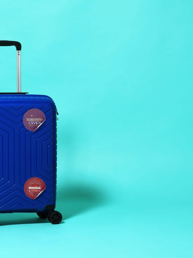 12 Canada cities as glitter pin badges - transparent PNG + printable A4 sheet. Print at home on sticker paper, cut, and use for journals, laptops, water bottles.Stickers are on a blue suitcase by Alter Standard.