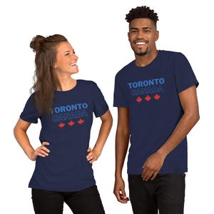 Op de afbeelding: Een marineblauw t-shirt met korte mouwen voorzien van de tekst TORONTO CANADA in blauwe en witte letters, vergezeld door drie kleine rode esdoornbladpictogrammen die horizontaal onder de tekst op de voorzijde van het kledingstuk zijn geplaatst.