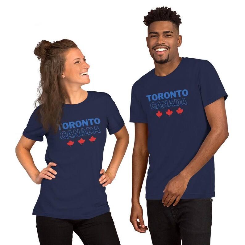 Op de afbeelding: Een marineblauw t-shirt met korte mouwen voorzien van de tekst TORONTO CANADA in blauwe en witte letters, vergezeld door drie kleine rode esdoornbladpictogrammen die horizontaal onder de tekst op de voorzijde van het kledingstuk zijn geplaatst.