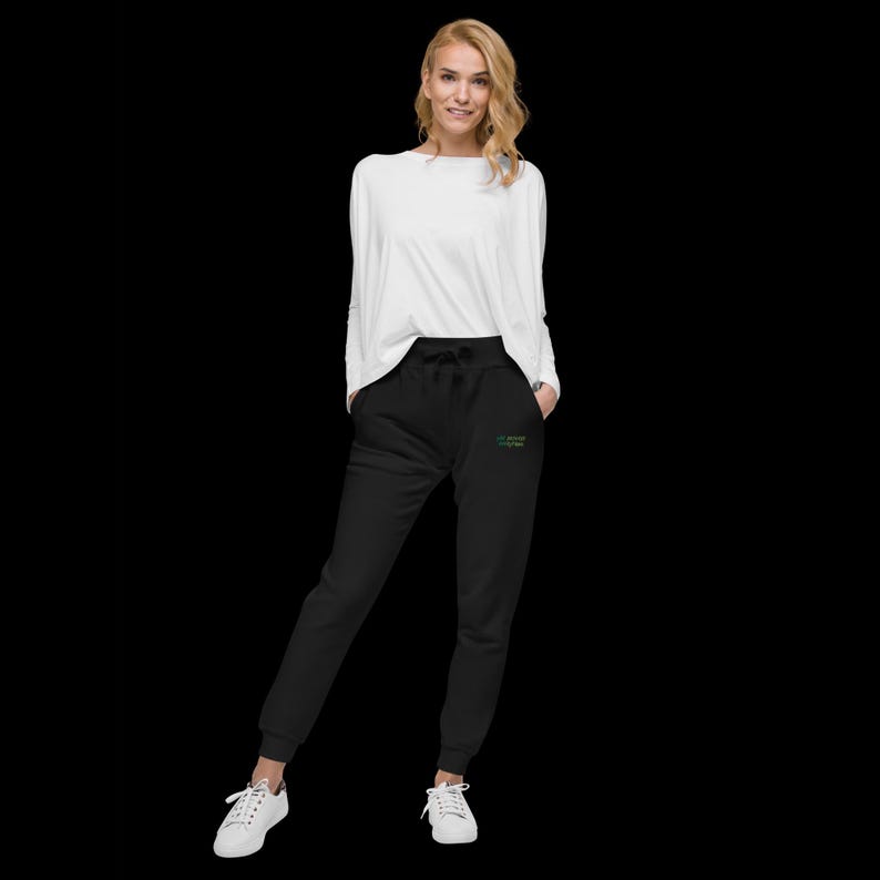May include: A long-sleeved white crewneck shirt paired with black joggers featuring a drawstring waist and ribbed cuffs. The joggers include a small, embroidered green and yellow text graphic on the upper left thigh area, worn with white trainers.