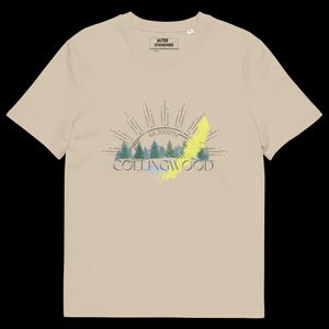 Pode incluir: Uma t-shirt bege de manga curta com um gr&aacute;fico de uma floresta, um sol nascente e o texto "COLLINGWOOD" com "44.5000&deg;N" acima. O design inclui um detalhe de pincelada amarela. A etiqueta interna diz "ALTER STANDARD".
