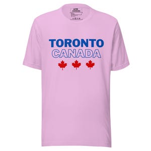 Op de afbeelding: Een lichtroze T-shirt met korte mouwen met de blauwe tekst TORONTO en de blauw omlijnde tekst CANADA, gevolgd door drie rode esdoornbladpictogrammen in het midden op de borst. Het label in de nek bevat de tekst ALTER STANDARD.