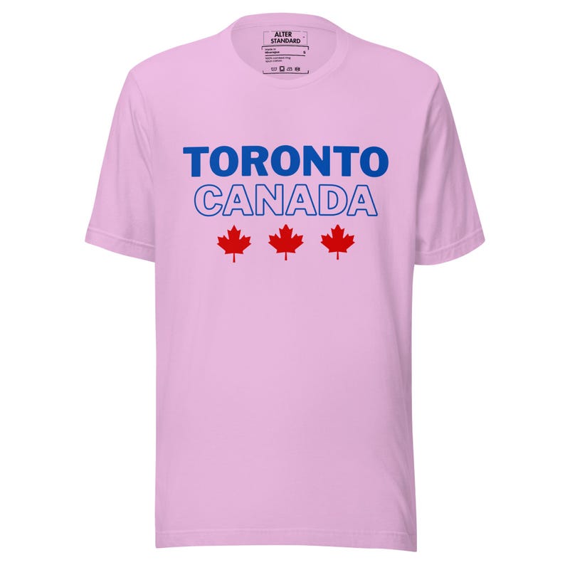 Op de afbeelding: Een lichtroze T-shirt met korte mouwen met de blauwe tekst TORONTO en de blauw omlijnde tekst CANADA, gevolgd door drie rode esdoornbladpictogrammen in het midden op de borst. Het label in de nek bevat de tekst ALTER STANDARD.