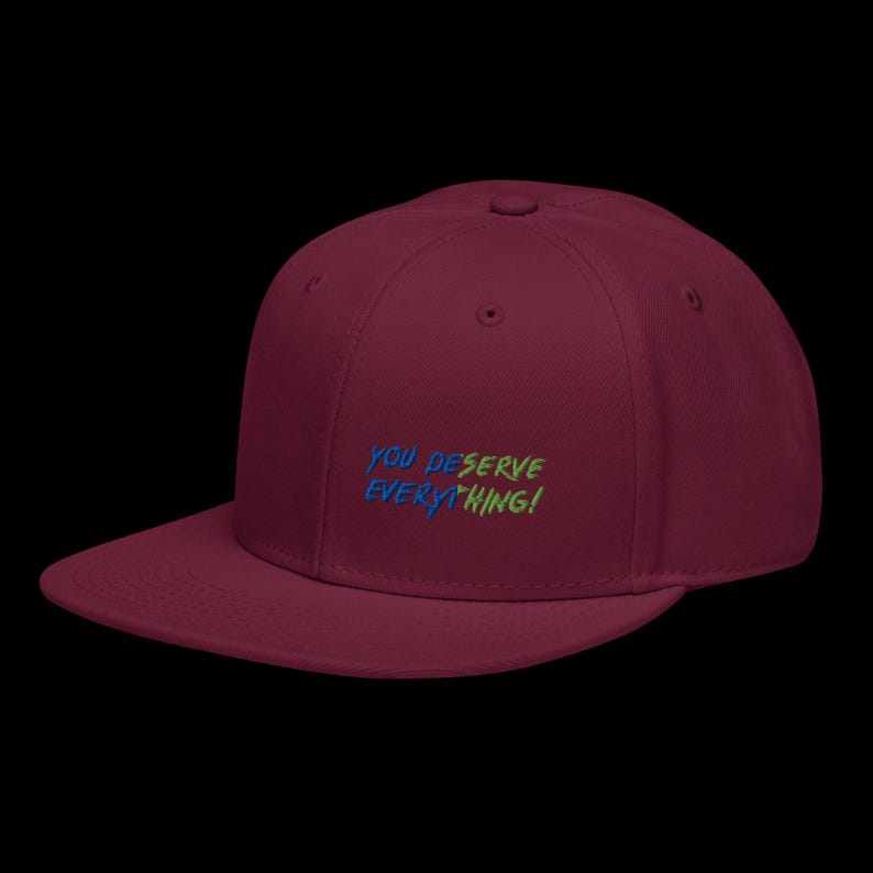 May include: A maroon baseball cap with a flat brim and embroidered text on the front that reads YOU DESERVE EVERYTHING! in alternating blue and green letters. The cap features a structured crown with a top button and stitched eyelets.