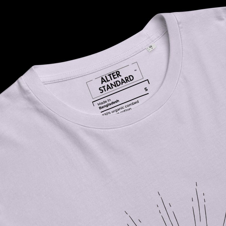 Pode incluir: Uma t-shirt lavanda claro com gola redonda e uma etiqueta interna impressa que diz ALTER STANDARD, Made in Bangladesh e 100% organic combed cotton. A pe&ccedil;a inclui uma etiqueta de tamanho S e um gr&aacute;fico parcial de linhas pretas.