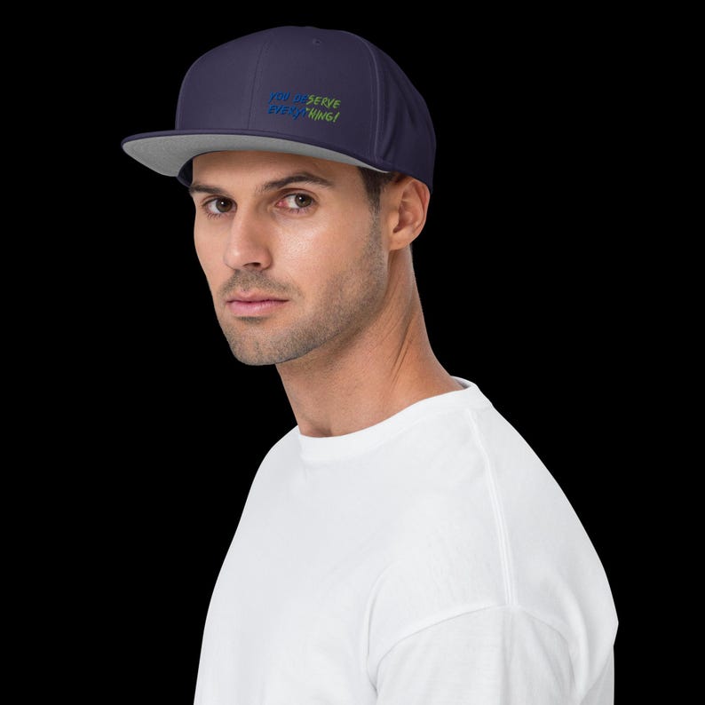 May include: A navy blue baseball cap with a flat brim and a light grey underside. The front of the cap features embroidered text in blue and green that reads "YOU DESERVE EVERYTHING!" in a clean, sans-serif font style on the front panel.