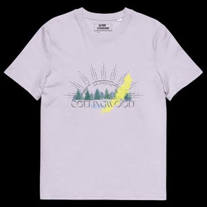 Pode incluir: Uma t-shirt de manga curta em roxo claro com um gr&aacute;fico de pinheiros verdes, um sol radiante e o texto "COLLINGWOOD" com "44.5000&deg;N" impresso acima. O design inclui um detalhe de pincelada amarela atr&aacute;s do texto e das &aacute;rvores.