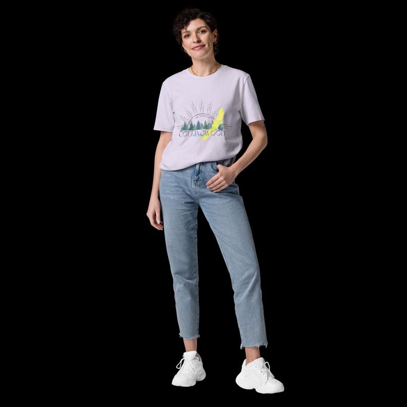 Pode incluir: Uma t-shirt de manga curta em lil&aacute;s claro com um gr&aacute;fico de uma floresta, um sol nascente e o texto COLLINGWOOD, combinada com cal&ccedil;as de ganga azul claro com bainha desfiada e sapatilhas brancas com sola grossa e atacadores.