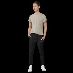 May include: A beige short-sleeved crew neck t-shirt paired with black joggers featuring a small green embroidered text detail on the left thigh and white athletic trainers. The outfit is presented against a solid black background.
