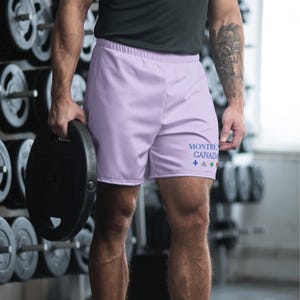May include: Light purple athletic shorts featuring the text MONTR&Eacute;AL CANADA in blue lettering on the lower left leg, accompanied by a small blue fleur-de-lis symbol and three additional small colorful icons arranged in a horizontal row.