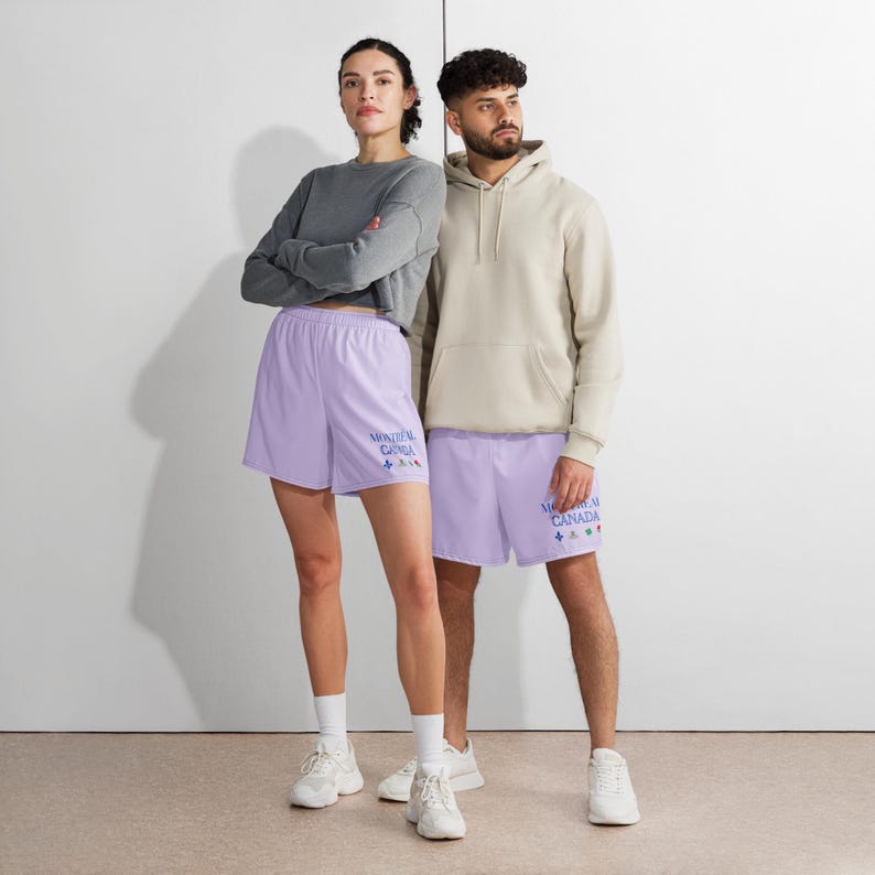 May include: A pair of light purple athletic shorts by Alter Standard featuring the blue text Montreal Canada and small colorful icons on the left leg. The shorts have an elastic waistband and are worn with white sneakers, a gray long-sleeve top, and a beige hoodie.