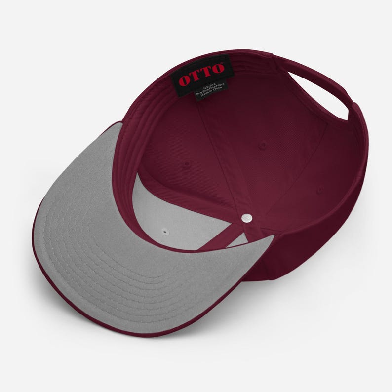May include: A maroon baseball cap viewed from the inside, featuring a grey underside on the brim and a black interior tag with the red text OTTO. The cap has a structured crown with a central silver button and an adjustable strap at the back.