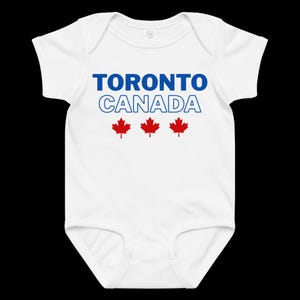以下が含まれることがあります： 青い文字で「TORONTO」、青い輪郭の文字で「CANADA」とプリントされた白い半袖のベビー用ボディスーツ。テキストの下には3つの赤いカエデの葉のアイコンが中央に配置されています。丸首で、股下にはスナップボタンが付いています。