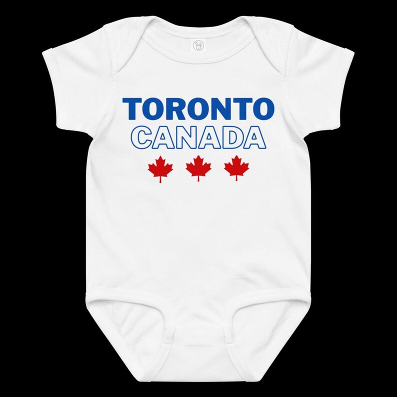 以下が含まれることがあります： 青い文字で「TORONTO」、青い輪郭の文字で「CANADA」とプリントされた白い半袖のベビー用ボディスーツ。テキストの下には3つの赤いカエデの葉のアイコンが中央に配置されています。丸首で、股下にはスナップボタンが付いています。