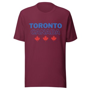 Op de afbeelding: Een bordeauxrood t-shirt met korte mouwen voorzien van de tekst TORONTO in blauwe letters, CANADA in blauwe omtrekletters en drie rode esdoornbladpictogrammen in een horizontale rij onder de tekst op de voorzijde van het kledingstuk.