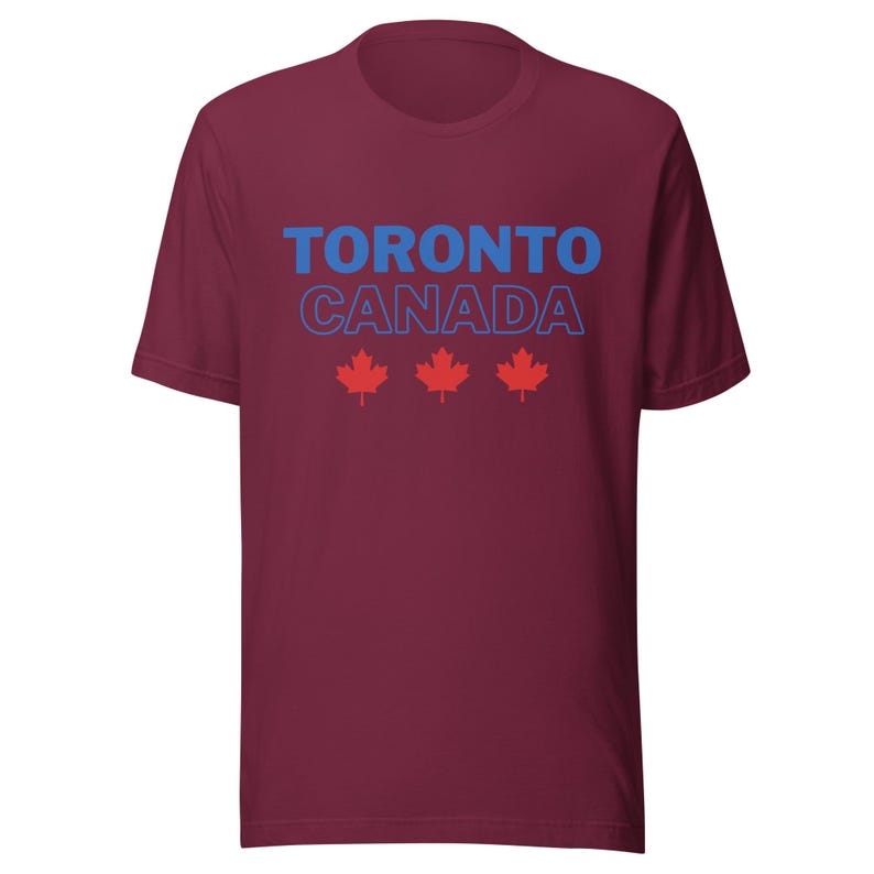 Op de afbeelding: Een bordeauxrood t-shirt met korte mouwen voorzien van de tekst TORONTO in blauwe letters, CANADA in blauwe omtrekletters en drie rode esdoornbladpictogrammen in een horizontale rij onder de tekst op de voorzijde van het kledingstuk.