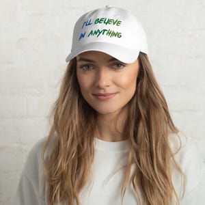 May include: A white baseball cap featuring the text I'LL BELIEVE IN ANYTHING embroidered in a gradient of blue and green colours across the front panel. The cap has a structured crown, a curved brim, and a button on the top centre of the headwear.