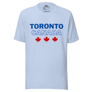 Op de afbeelding: Een lichtblauw t-shirt met korte mouwen voorzien van de tekst TORONTO in effen blauw, CANADA in blauwe omlijning en drie rode esdoornbladiconen in het midden eronder. Het label in de nek toont de merknaam ALTER STANDARD en wasinstructies.