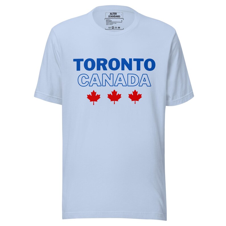 Op de afbeelding: Een lichtblauw t-shirt met korte mouwen voorzien van de tekst TORONTO in effen blauw, CANADA in blauwe omlijning en drie rode esdoornbladiconen in het midden eronder. Het label in de nek toont de merknaam ALTER STANDARD en wasinstructies.