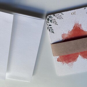 May include: A stack of white envelopes next to a stack of rectangular cards bound by a brown paper band. The cards feature a watercolour-style red heart design, black botanical illustrations in the corners, and the number 20 printed at the top.