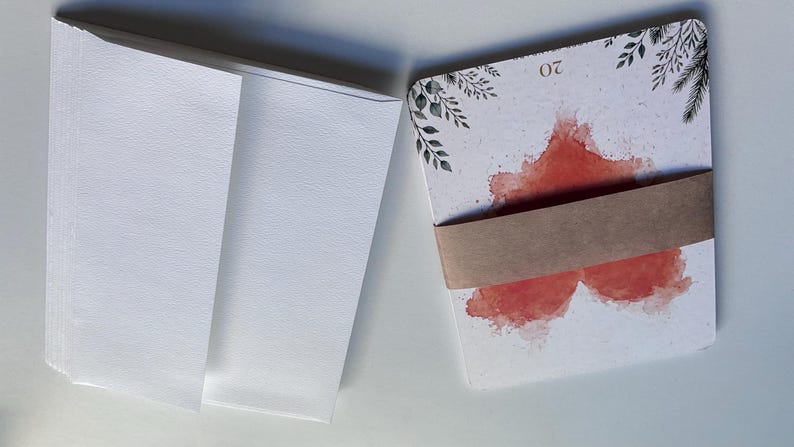 May include: A stack of white envelopes next to a stack of rectangular cards bound by a brown paper band. The cards feature a watercolour-style red heart design, black botanical illustrations in the corners, and the number 20 printed at the top.