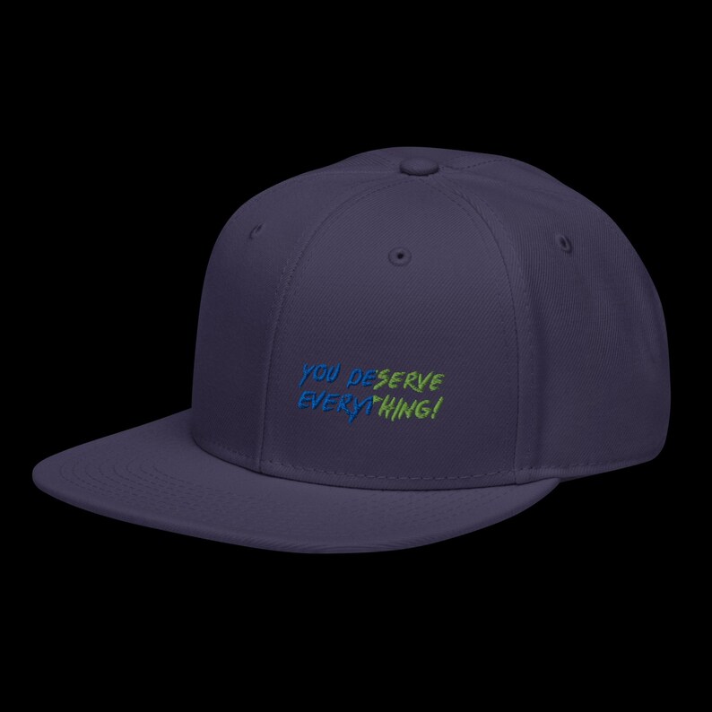 May include: A dark purple baseball cap with a flat brim and a structured crown. The front features embroidered text that reads YOU DESERVE EVERYTHING! in a split colour design, with the first half in blue and the second half in lime green.
