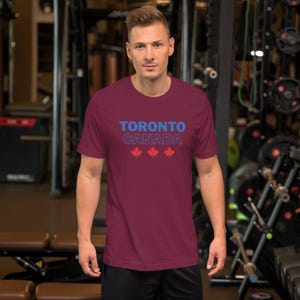 Op de afbeelding: Een bordeauxrood t-shirt met korte mouwen, voorzien van de tekst TORONTO in het blauw, CANADA in een blauwe omlijning en drie rode esdoornblad-iconen in het midden op de borst. Het shirt heeft een ronde hals en een standaard pasvorm.