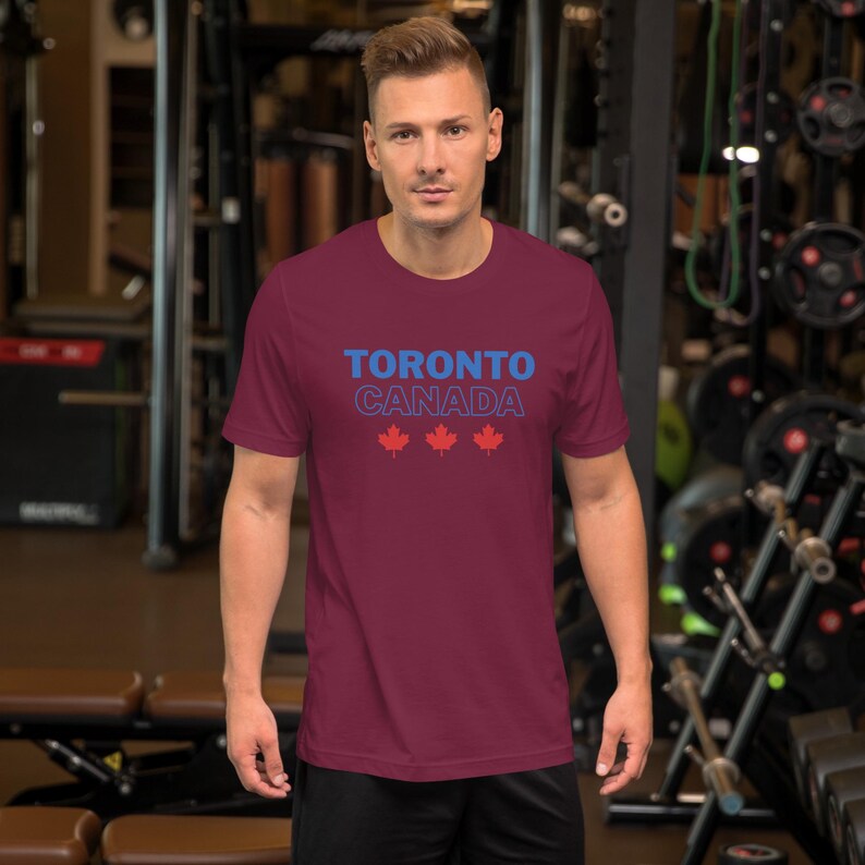 Op de afbeelding: Een bordeauxrood t-shirt met korte mouwen, voorzien van de tekst TORONTO in het blauw, CANADA in een blauwe omlijning en drie rode esdoornblad-iconen in het midden op de borst. Het shirt heeft een ronde hals en een standaard pasvorm.