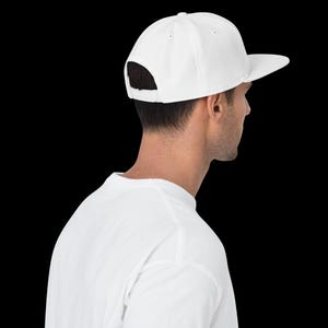 May include: A solid white baseball cap with a structured crown and a flat brim. The cap features an adjustable snapback closure at the back, providing a customisable fit. The design is clean and simple, showcasing a plain white fabric construction.