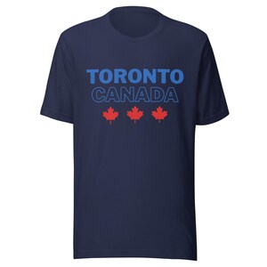 Op de afbeelding: Een marineblauw t-shirt met korte mouwen voorzien van de tekst TORONTO in effen blauwe letters boven het woord CANADA in een blauw omlijnd lettertype. Onder de tekst staan drie rode esdoornblad-iconen horizontaal op de borst.
