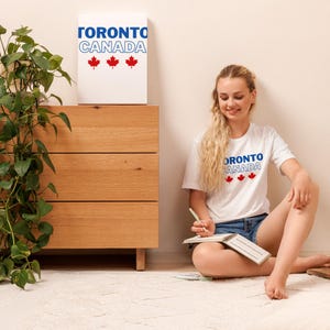 Op de afbeelding: Een wit T-shirt met korte mouwen voorzien van de blauwe tekst TORONTO, de blauw omlijnde tekst CANADA en drie rode esdoornblad-iconen. Hetzelfde grafische ontwerp staat op een wit rechthoekig wandpaneel op een houten ladekast.