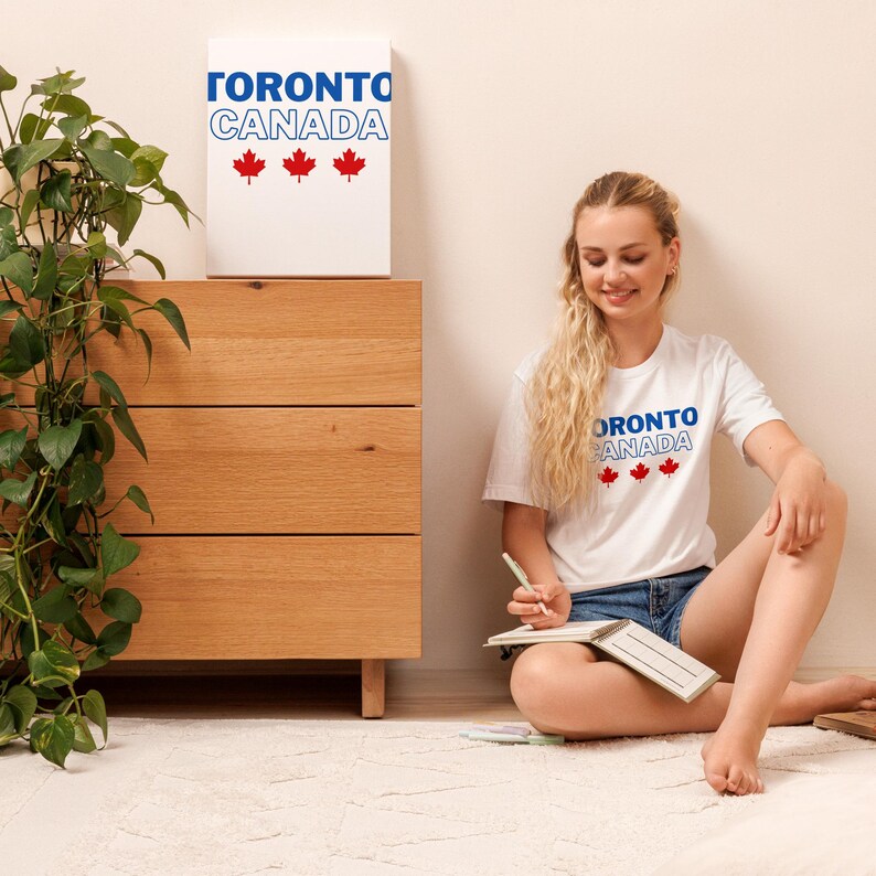 Op de afbeelding: Een wit T-shirt met korte mouwen voorzien van de blauwe tekst TORONTO, de blauw omlijnde tekst CANADA en drie rode esdoornblad-iconen. Hetzelfde grafische ontwerp staat op een wit rechthoekig wandpaneel op een houten ladekast.