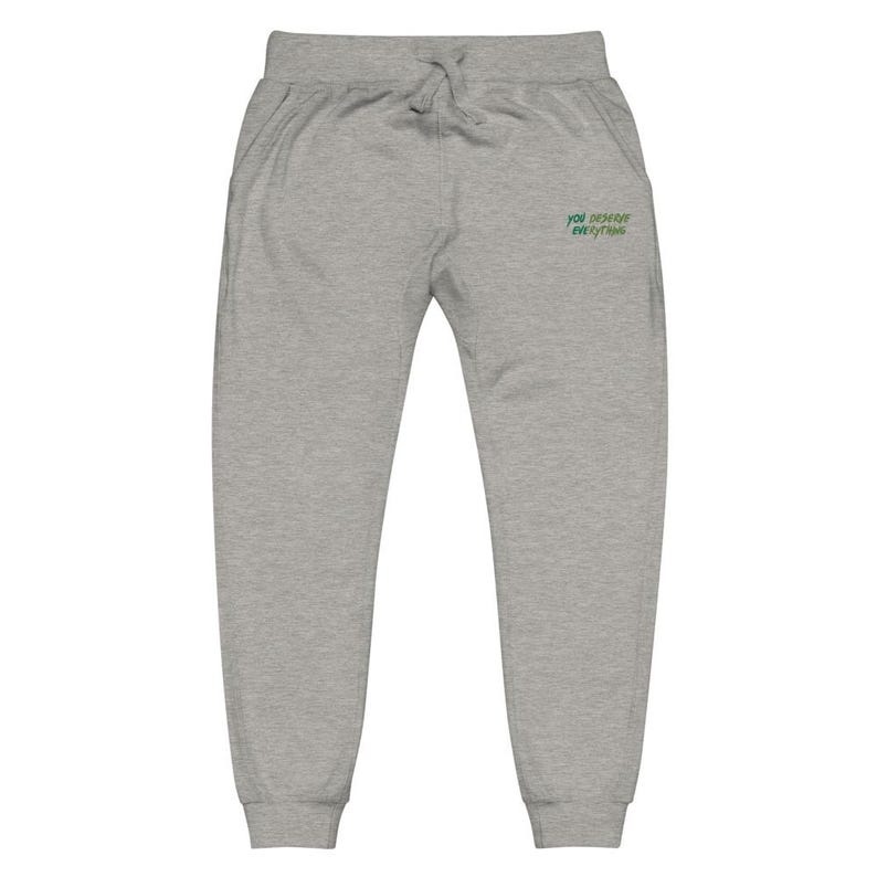 May include: Light grey sweatpants featuring an elastic waistband with a drawstring and ribbed cuffs at the ankles. The front right side displays the embroidered green text "YOU DESERVE EVERYTHING" in a stylised, slightly slanted font.