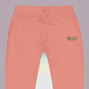 May include: Peach-coloured sweatpants featuring an elastic waistband with a matching drawstring. The right side displays the text "YOU DESERVE EVERYTHING" printed in a dark green, handwritten-style font against the solid peach fabric.