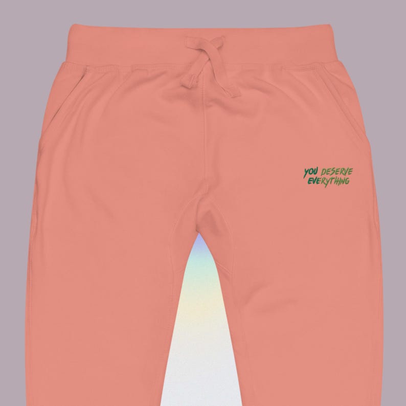 May include: Peach-coloured sweatpants featuring an elastic waistband with a matching drawstring. The right side displays the text "YOU DESERVE EVERYTHING" printed in a dark green, handwritten-style font against the solid peach fabric.