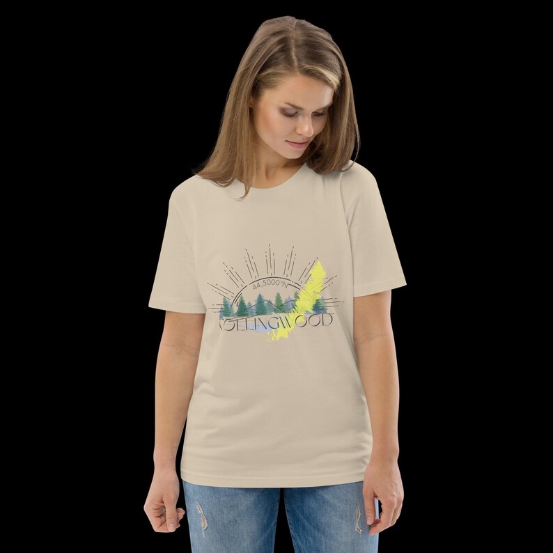 Pode incluir: Uma t-shirt de manga curta bege com uma impress&atilde;o gr&aacute;fica de uma paisagem florestal com um design de raios de sol por cima. O texto "NOTTINGWOOD" est&aacute; impresso em letras pretas no centro, acompanhado por um detalhe de pincelada amarela.