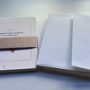 May include: A white rectangular card set with a brown paper band featuring the text Small Luck Arrives and Japanese characters. A small red ladybug illustration is visible below the text. Beside it sits a stack of white envelopes.