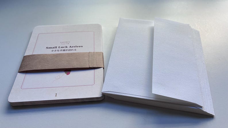 May include: A white rectangular card set with a brown paper band featuring the text Small Luck Arrives and Japanese characters. A small red ladybug illustration is visible below the text. Beside it sits a stack of white envelopes.