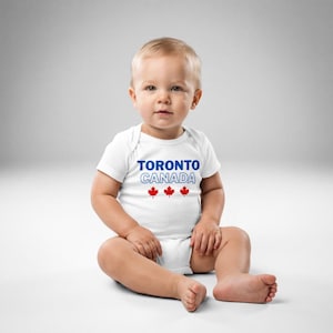以下が含まれることがあります： 胸の中央に青い文字でTORONTO、白い縁取りの文字でCANADA、その下に3つの小さな赤いカエデの葉のアイコンがプリントされた、白い半袖のベビー用ボディスーツ。丸首で、股下にスナップボタンが付いています。