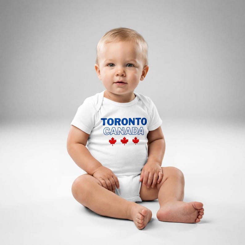 以下が含まれることがあります： 胸の中央に青い文字でTORONTO、白い縁取りの文字でCANADA、その下に3つの小さな赤いカエデの葉のアイコンがプリントされた、白い半袖のベビー用ボディスーツ。丸首で、股下にスナップボタンが付いています。