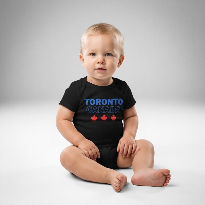 以下が含まれることがあります： 胸元に青い文字でTORONTOとCANADAがプリントされ、その下に3つの小さな赤いカエデの葉のアイコンが横一列に並んだ、黒い半袖のベビー用ボディスーツ。