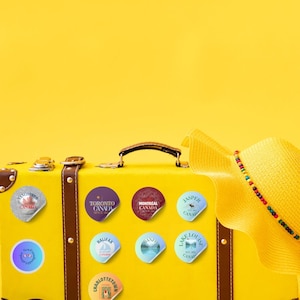 12 Canada cities as glitter pin badges - transparent PNG + printable A4 sheet. Print at home on sticker paper, cut, and use for suitcases, journals, laptops, water bottles. Stickers are on a yellow suitcase. Designed by Alter Standard