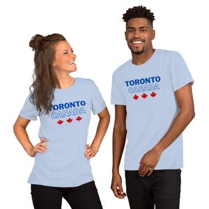 Op de afbeelding: Een lichtblauw t-shirt met korte mouwen voorzien van de tekst TORONTO CANADA in blauwe en witte omtrekletters, vergezeld door drie kleine rode esdoornbladpictogrammen die horizontaal onder de tekst op de voorzijde zijn geplaatst.
