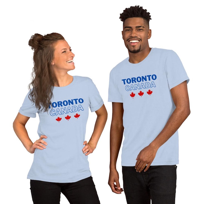 Op de afbeelding: Een lichtblauw t-shirt met korte mouwen voorzien van de tekst TORONTO CANADA in blauwe en witte omtrekletters, vergezeld door drie kleine rode esdoornbladpictogrammen die horizontaal onder de tekst op de voorzijde zijn geplaatst.