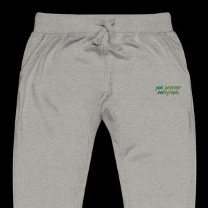 May include: Light grey sweatpants featuring an elastic waistband with a drawstring. The right side displays the embroidered green text "YOU DESERVE EVERYTHING" in a stylised, slightly jagged font. The garment has a simple, clean design.