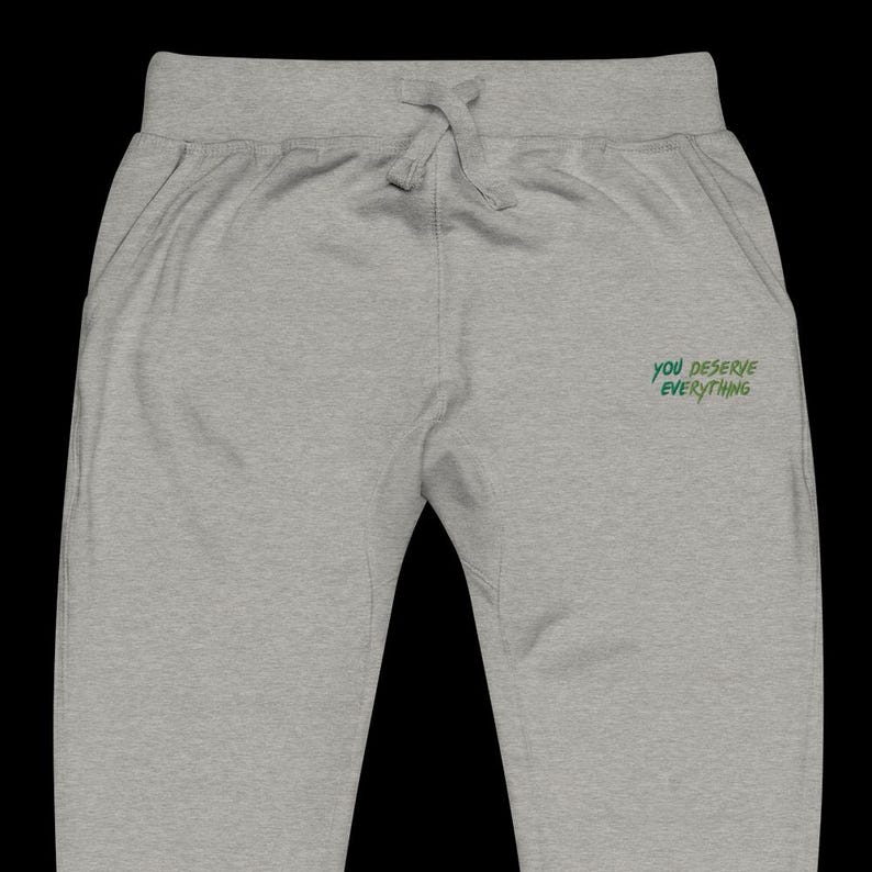 May include: Light grey sweatpants featuring an elastic waistband with a drawstring. The right side displays the embroidered green text "YOU DESERVE EVERYTHING" in a stylised, slightly jagged font. The garment has a simple, clean design.